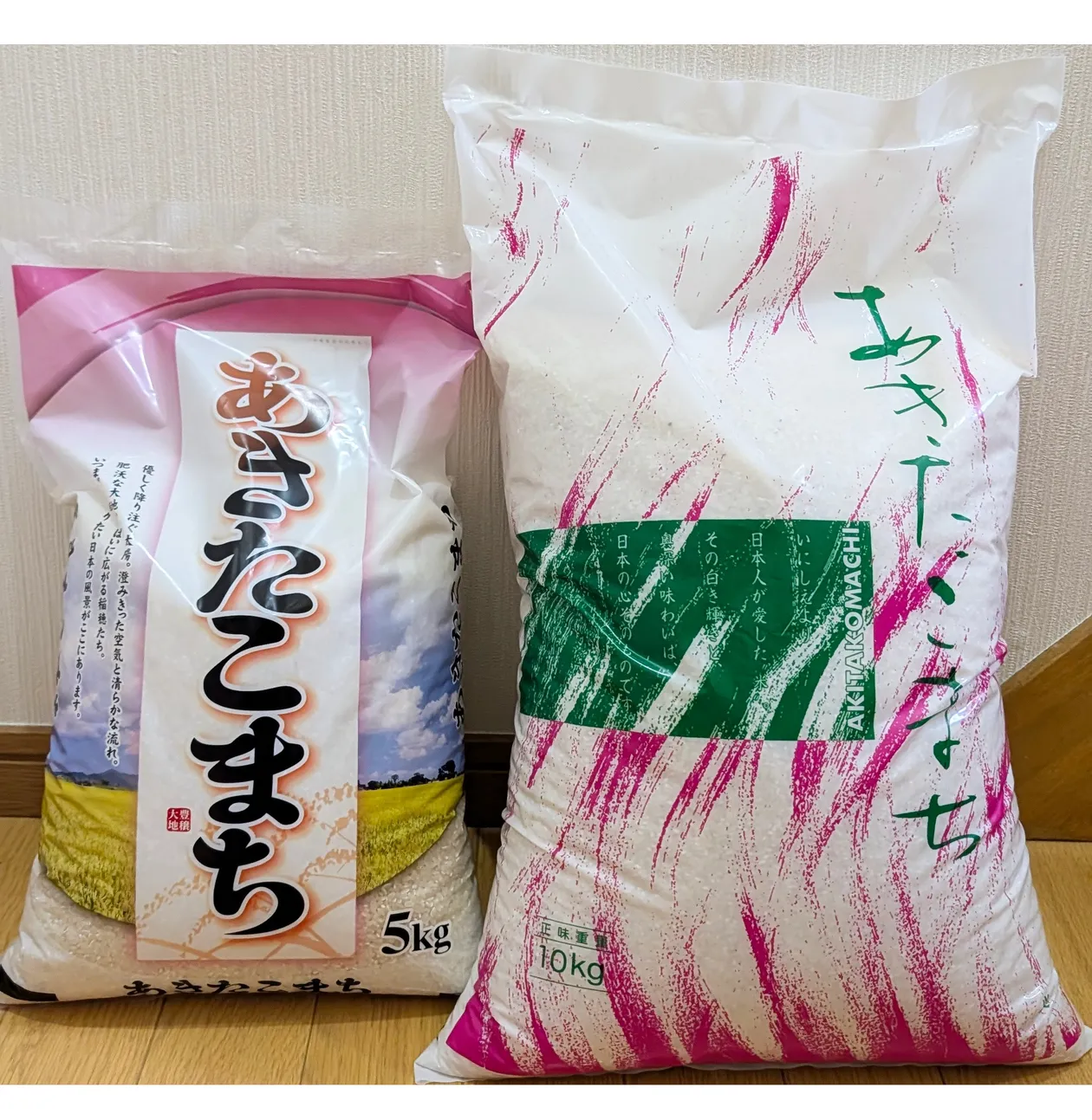 白い壁を背に立てて並べた、あきたこまちの5kg袋(左・ピンク基調のパッケージ)と10kg袋(右・白地にピンクの筆跡と緑の帯)。